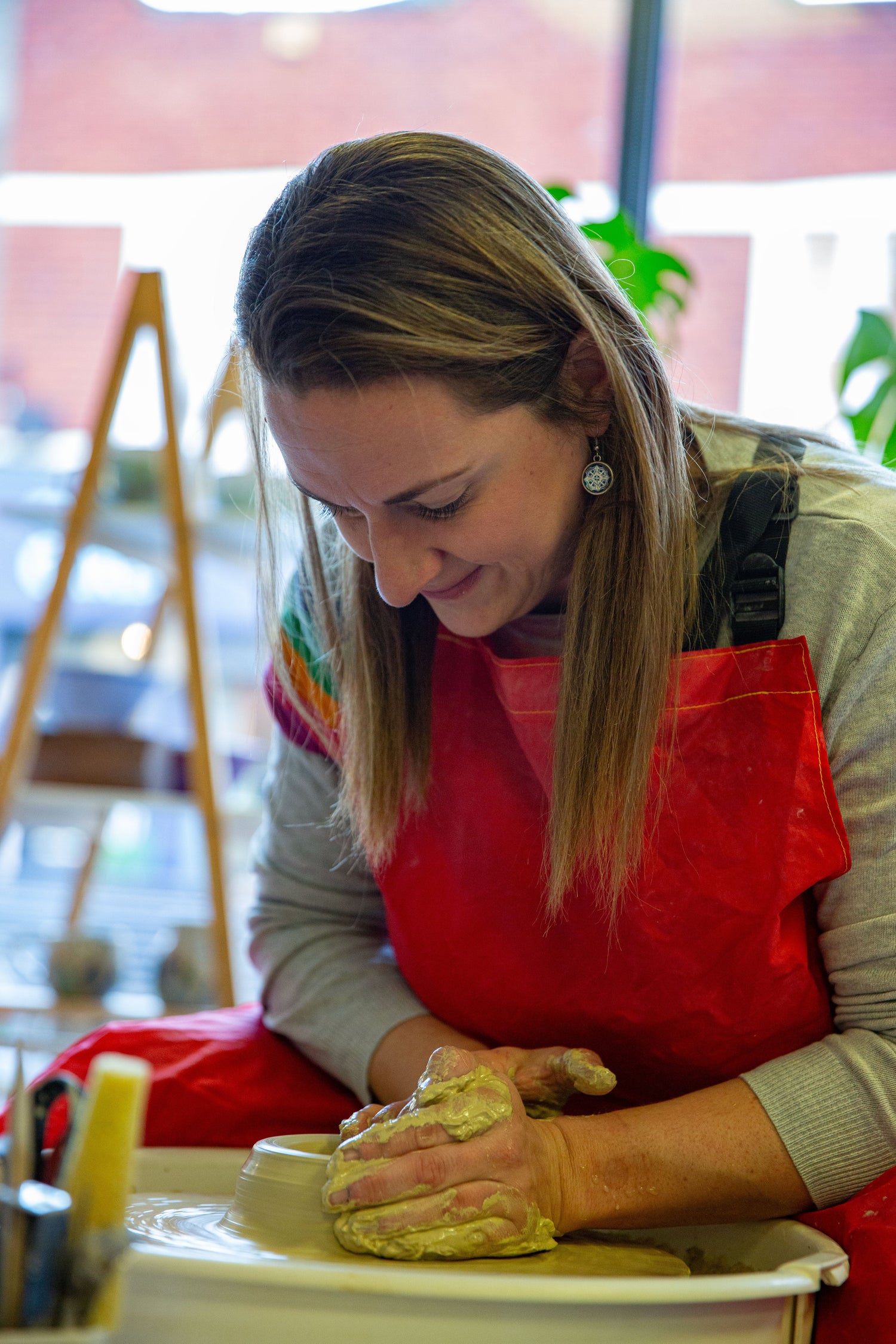 Woman with straight brown hair sits at a pottery wheel focusing on the clay that she is throwing with.